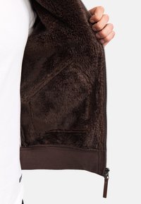 Chaqueta marrón con cremallera y un interior suave y aterciopelado. Presenta una cremallera oscura y un dobladillo marrón sólido, ofreciendo una apariencia acogedora y texturada.