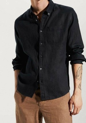 Camicia button-up in lino di un blu scuro, con maniche lunghe, taschino sul petto e bottoni visibili, abbinata a pantaloni color marrone chiaro.
