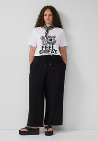 Weißes T-Shirt mit schwarzem Text und Grafik, kombiniert mit lockeren schwarzen Hosen. Schwarze Plateausandalen mit sichtbaren Riemen am Knöchel.