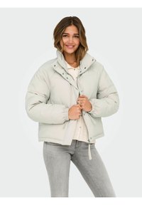 Chaqueta acolchada gris claro con cuello alto, cierre de cremallera y broches, puños elásticos y diseño corto, llevada sobre una camiseta crema y vaqueros grises.