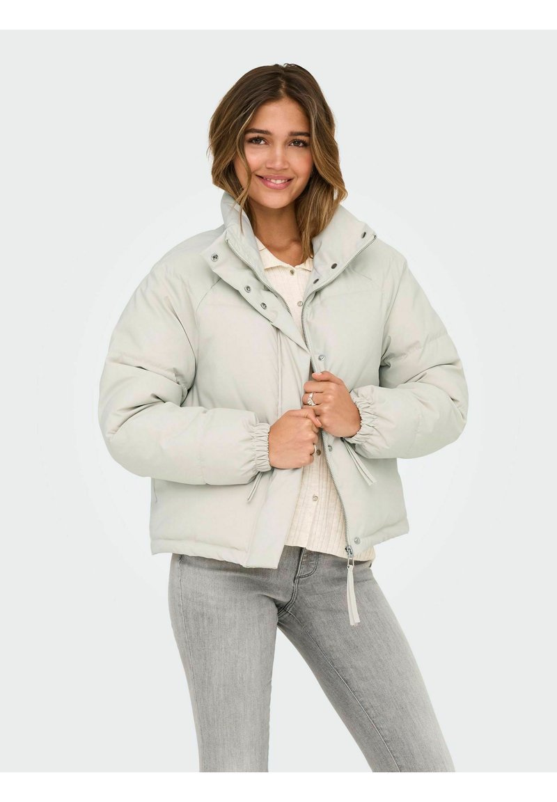 Chaqueta acolchada gris claro con cuello alto, cierre de cremallera y broches, puños elásticos y diseño corto, llevada sobre una camiseta crema y vaqueros grises.