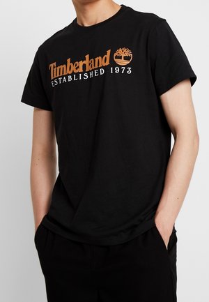Maglietta nera in cotone con logo "Timberland" in arancione e bianco, con la data di fondazione "1973" e l'emblema dell'albero. Maniche corte, design con scollo rotondo.