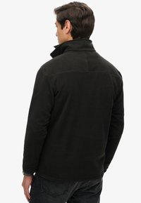 Chaqueta de forro polar negra con cuello alto, que presenta una textura suave y un detalle sutil de costuras a lo largo de los hombros y la espalda.