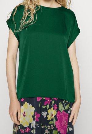 Femme portant un haut à manches courtes vert foncé associé à une jupe bleu marine ornée de grands motifs floraux jaunes, roses et blancs.