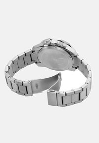 Ura iz nerjavečega jekla z verižico, srebrne barve, okroglo ohišje, dvema gumboma in zaponko. Gladka, polirana površina z vgraviranim logotipom.