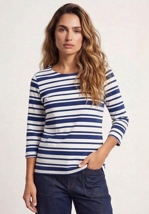 Femme aux longs cheveux ondulés portant un haut à manches longues rayé bleu et blanc et un pantalon bleu foncé, debout avec une main dans la poche.