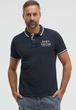 Mann trägt ein marineblaues Polohemd mit weißen und blauen Abschlüssen, bestickten Text auf der Brust und graue Hosen, steht mit den Händen in den Taschen.