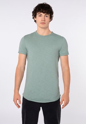 Mann mit lockigem dunklem Haar, der ein hellgrünes T-Shirt mit Rundhalsausschnitt und kurzen Ärmeln sowie schwarze Hose trägt, steht vor einem einfarbigen Hintergrund.