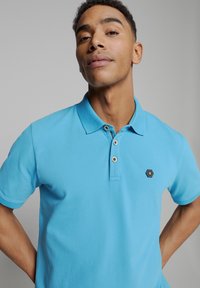 Lichtblauw poloshirt met een kraag, drie knopen en een hexagonaal logo op de borst. Zacht, textuur katoen.