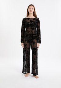 Woman stands barefoot wearing black sheer floral long-sleeve top and matching wide-leg pants over black undergarments, white background.