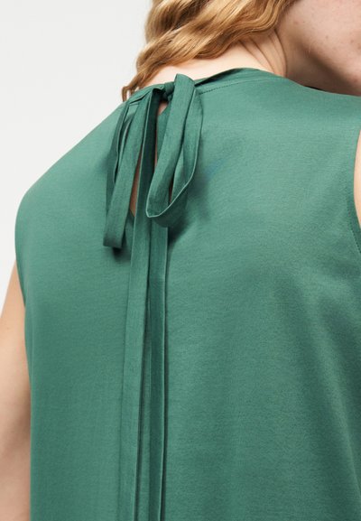 Πράσινο αμάνικο τοπ με υφασμάτινο δέσιμο στο πίσω μέρος του λαιμού, φορεμένο από άτομο με ανοιχτό χρώμα δέρματος και ανοιχτό καφέ μαλλιά.