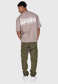 Braunes, übergroßes T-Shirt mit weißem grafischem Text auf dem Rücken, kombiniert mit olivgrünen Cargo-Hosen mit Seitentaschen und weißen Sneakers.