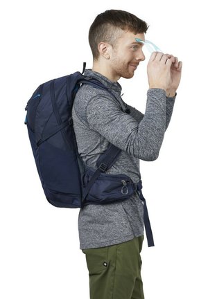 Young man wearing gray long-sleeve shirt and green pants adjusts blue safety glasses, carrying a large navy blue hiking backpack.