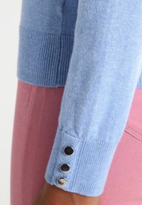 Ljusblå tröja med ribbad manschett och tre dekorativa knappar, i par med ett rosa plagg i bakgrunden.