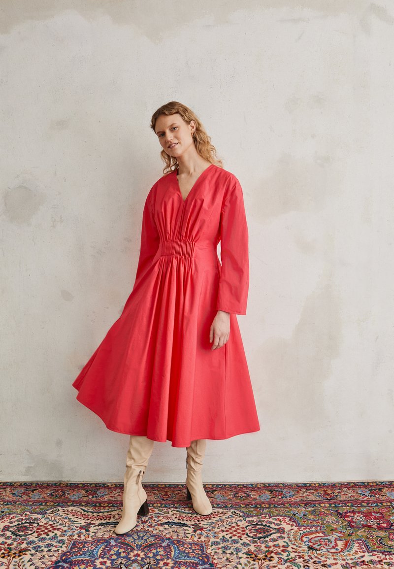 Vestido de algodão vermelho com decote em V e mangas longas, apresentando detalhes franzidos na cintura e uma saia rodada, combinado com botas bege até ao joelho.