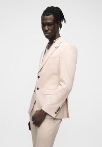 Homme aux cheveux tressés portant un costume beige clair et une chemise gris clair, debout devant un fond blanc uni.