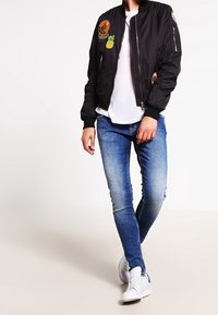 Svart bomberjacka med broderade tygmärken, vit t-shirt, blekta blå skinny jeans och vita sneakers. Modellen står med ena handen på höften.