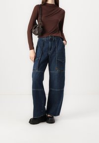 Lösa, mörkblå denim cargo-byxor med flera fickor och kontrasterande sömmar. Matchat med en brun långärmad topp och svarta skor.