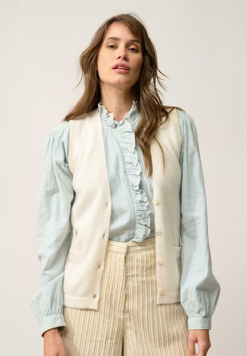 Kremowa kamizelka z pięcioma guzikami na jasnoniebieskiej bluzce z falbanami. Beżowe spodnie w pionowe paski. Miękkie tekstury i warstwowy fason.