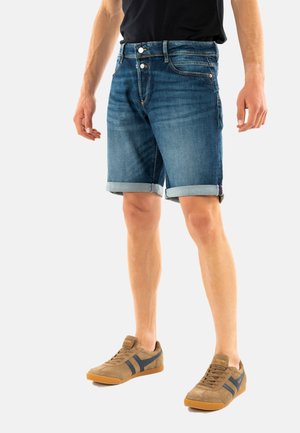 Shorts de mezclilla de color azul medio, con dobladillos enrollados, dos bolsillos frontales y cierre de cremallera. Combinados con zapatillas de ante marrón.