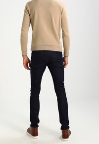 Jeans di denim blu scuro con una vestibilità slim, abbinati a un maglione beige. I jeans presentano tasche posteriori e dettagli di cuciture sottili.