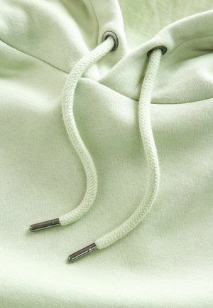 Svijetlozelena dukserica s mekim teksturama, vezicama s metalnim vrhovima i okruglom kapuljačom. Suptilni detalji šivanja su vidljivi.