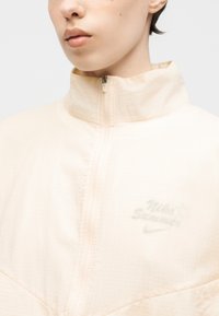 Zbliżenie osoby noszącej bladoróżową kurtkę zapinaną na zamek z wysokim kołnierzem i delikatnym logo Nike na piersi.