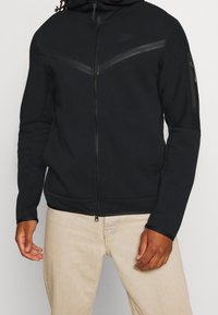 Man som bär en svart zip-up Nike hoodie med en bröstrand och ficka med blixtlås på vänster ärm, i kombination med beige byxor.