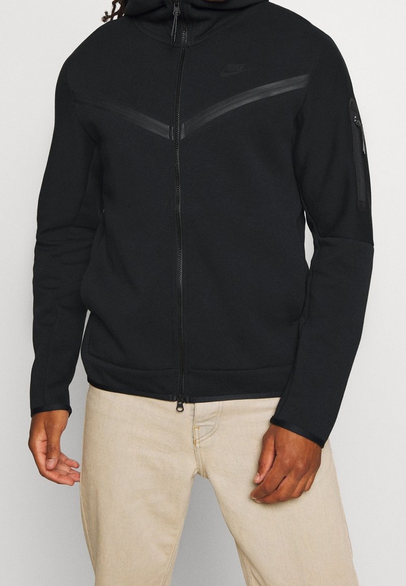 Man som bär en svart zip-up Nike hoodie med en bröstrand och ficka med blixtlås på vänster ärm, i kombination med beige byxor.