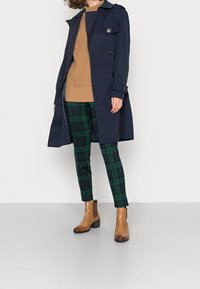 Marinblå trenchcoat, tan-stickad tröja, gröna rutiga byxor, bruna chelsea boots. Har knappar och bälte på rocken, strukturerad tröja.