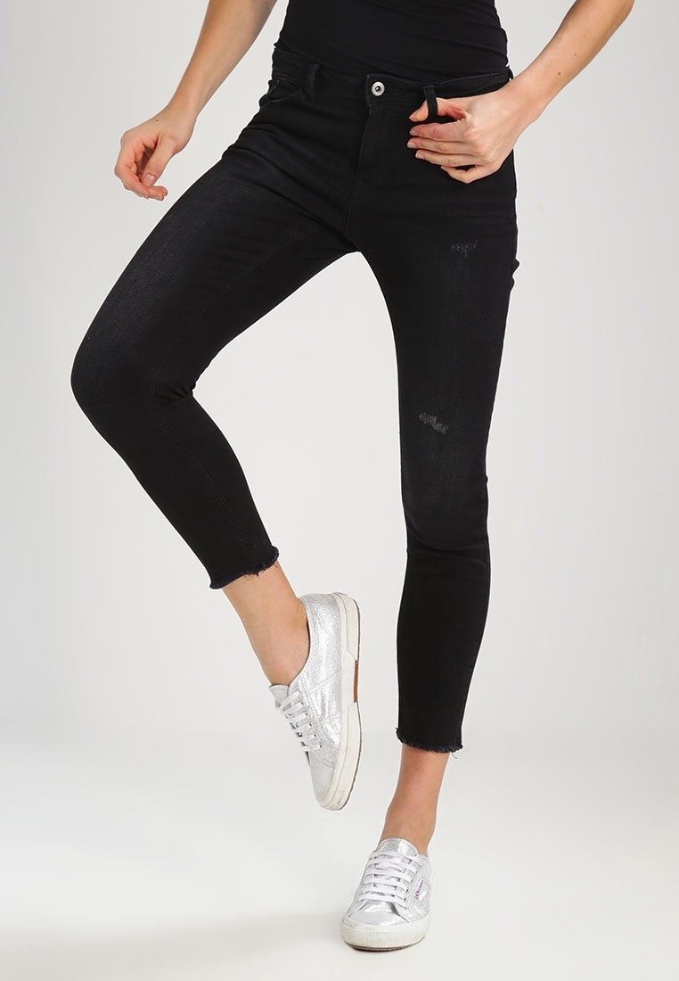 Jean skinny noir en denim à taille haute, légèrement décoloré, avec des distresses légères et des ourlets effilochés, portés avec des baskets argentées.