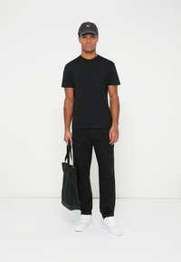 Melna kokvilnas T-kreklā, melnos biksēs, baltajās sporta apavās un tumšā cepurē ar logo. Tur rokās melna tote soma. Vienkārša, ikdieniška apģērba komplekts.