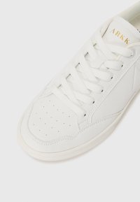 White leather sneaker with perforated toe, flat white laces, and a textured side panel. Features a logo in gold on the tongue.
