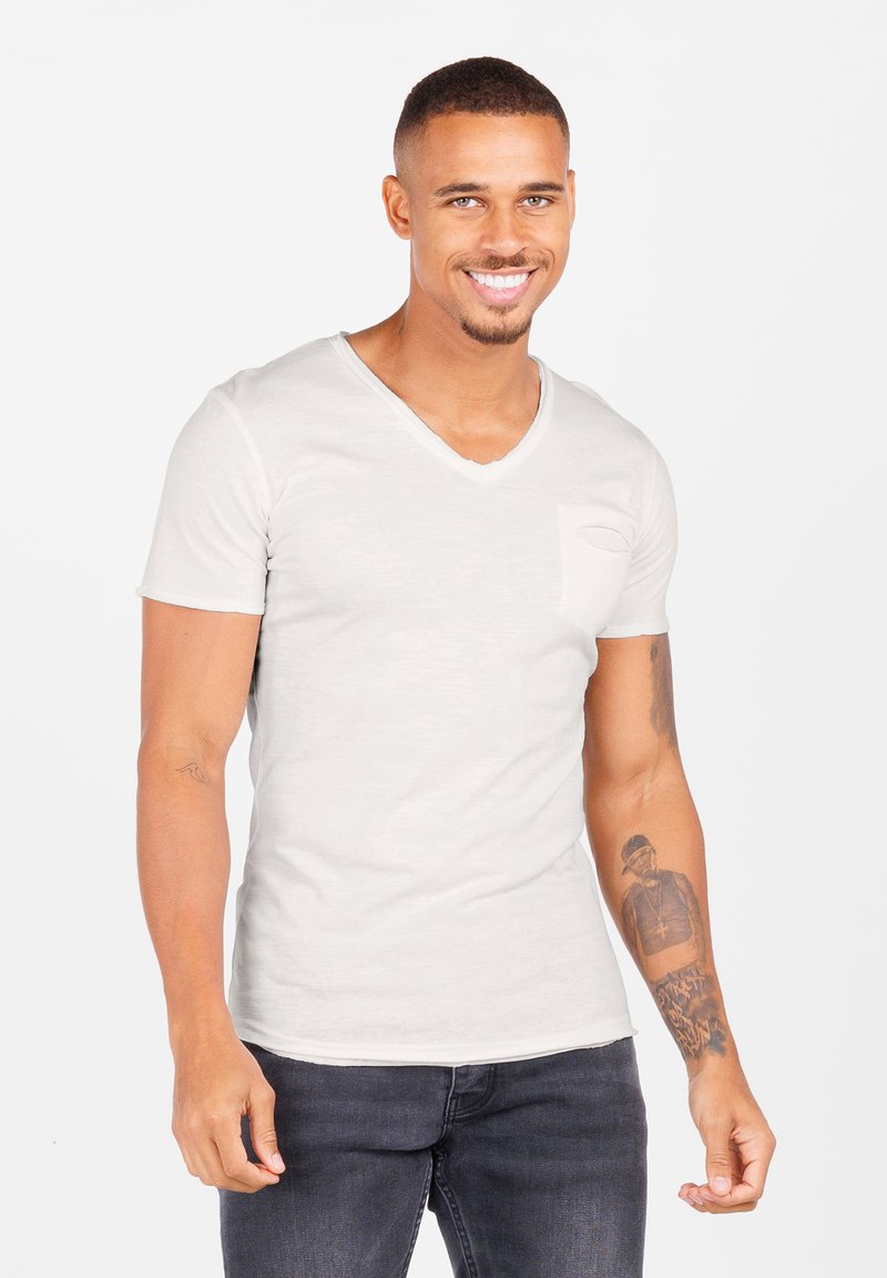 Camiseta de manga corta en tono gris claro con escote en V y un pequeño bolsillo en el pecho. Combina con jeans oscuros. La textura de la piel es visible en los brazos con detalles de tatuajes visibles.