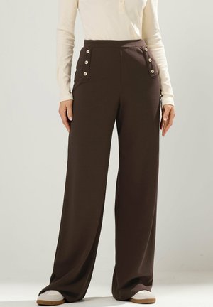 Kvinde iført mørkebrune, vidde bukser med høj talje og knapdetaljer, en cremefarvet langærmet top og hvide sko mod en ensfarvet baggrund.