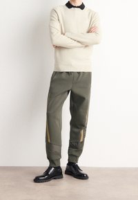 Suéter texturizado de un beige claro con un cuello negro, combinado con pantalones verdes oliva que tienen detalles dorados a los lados y zapatos negros con cordones.