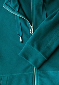 Teal zip-up hoodie med en texturerad tyg, framträdande dragkedja och ett mjukt innertyg, med en präglad logotyp på höger ärm.