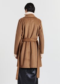 Cappotto in misto lana marrone con vita cinturata, colletto alto e polsini a coste color crema, visto da dietro su uno sfondo neutro.