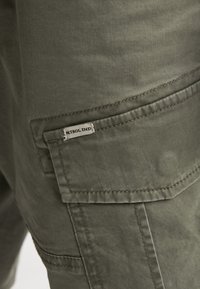 Pantalones cargo color verde oliva hechos de mezcla de algodón, con un gran bolsillo lateral y detalles acolchados. Etiqueta visible en el bolsillo.