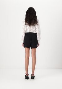 Femme aux longs cheveux bouclés portant un chemisier blanc en broderie anglaise, un short noir taille haute et des sandales noires à talons hauts, debout, tournée de dos.