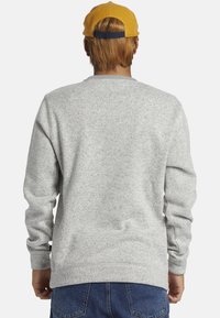 Grauer Hoodie aus strukturiertem Stoff, mit geripptem Halsausschnitt und Bündchen, versehen mit einem Etikett nahe dem unteren Saum. Die Rückansicht zeigt eine Schlichtheit im Design.