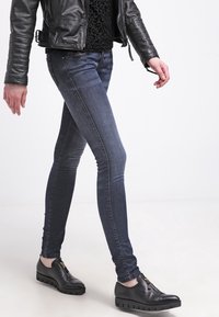 Jeans skinny de mezclilla en un tono oscuro con un ligero desgaste, combinados con una chaqueta de cuero negra y una blusa negra texturizada. Completados con zapatos negros sin cordones.