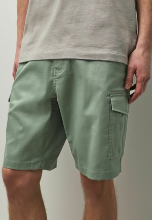 Mann trägt hellgrüne Cargoshorts und ein beiges T-Shirt, Hand ruht neben dem Oberschenkel, sichtbar von der Taille bis zu den Knien.
