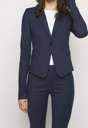 Femme portant un blazer bleu marine ajusté avec un bouton sur un top blanc et un pantalon bleu marine assorti, debout devant un fond uni.
