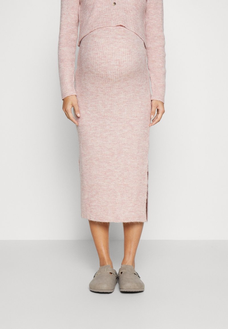 Gravid kvinna iklädd en ribbad rosa stickad klänning och gråa slip-on-skor, som står mot en enkel ljus bakgrund.