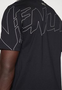 Camiseta negra de manga corta llevada por una persona de piel oscura, con grandes letras abstractas blancas contorneadas en la parte superior de la espalda y en las mangas.