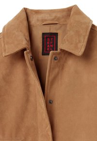 Braune Wildlederjacke mit kragen, Druckknöpfen und einer strukturierten Oberfläche. Im Kragen befindet sich ein rotes und schwarzes Etikett.