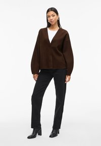 Ruda per didelį v-kaklą, ilgomis rankovėmis ir laisvu siluetu, derinama su juodomis tiesiomis kelnėmis ir kulkšnies batais.