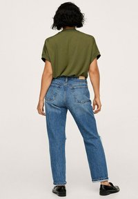 Oliwkowa bluzka z krótkim rękawem i wycięciem na plecach, w połączeniu z niebieskimi dżinsami o średnim stanie, charakteryzującymi się luźnym krojem i delikatnym przetarciem. Czarne buty.