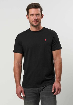 Homme portant un t-shirt noir uni à manches courtes avec un petit logo rouge sur la poitrine gauche et un jean gris foncé, debout devant un fond uni.
